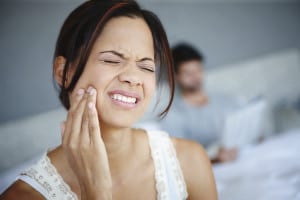 Shot of a woman sitting on the side of her bed with bad toothache with her boyfriend in the background